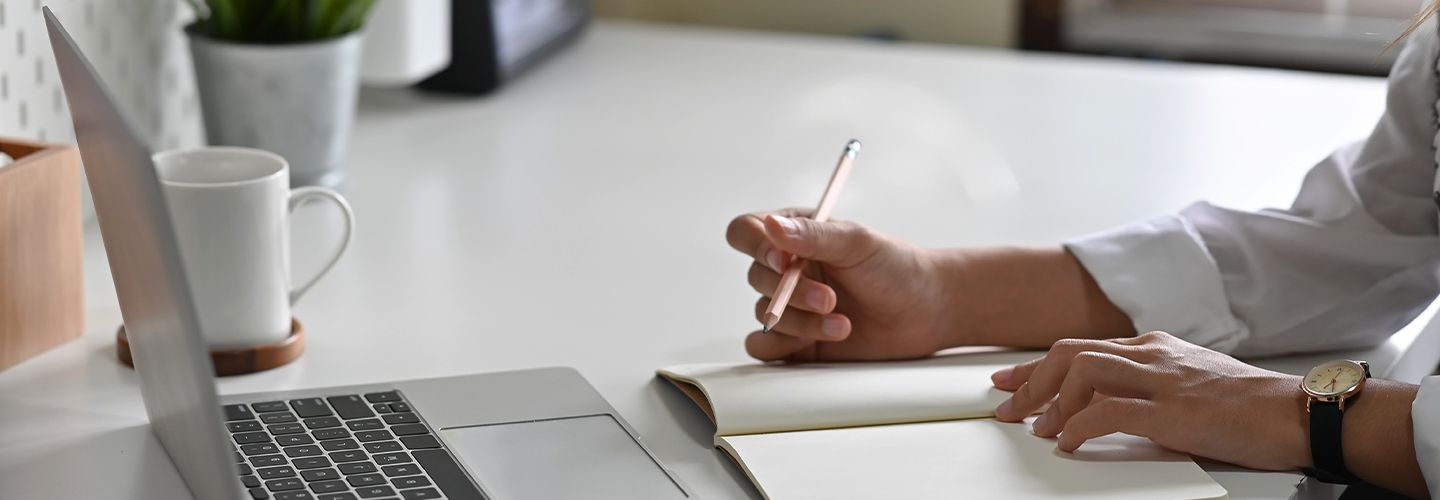 Kobieta patrzy w laptopa i zapisuje coś ołówkiem w notatniku.