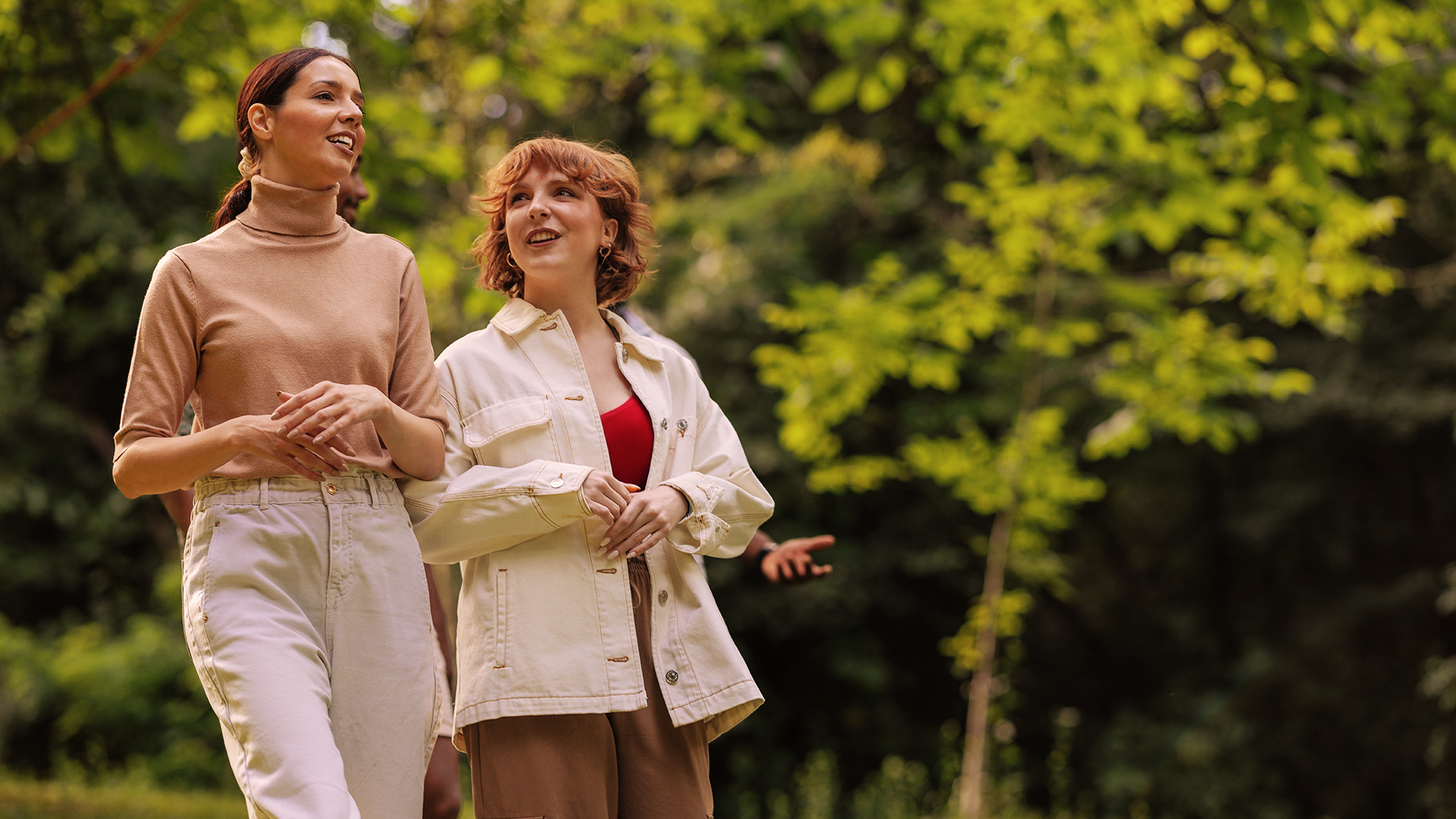 Dwie młode kobiety o jasnej karnacji spacerują latem po parku, rozmawiając z uśmiechem i ciesząc się słonecznym dniem.