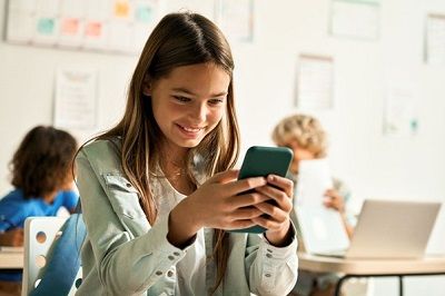 Uczennica szkoły podstawowej siedzi w ławce w klasie szkolnej i sprawdza cos w swoim telefonie.