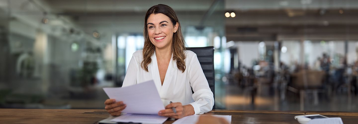 Uśmiechnięta kobieta siedzi przy biurku trzymając dokumenty.