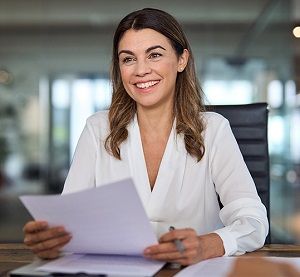 uśmiechnięta kobieta siedzi przy biurku trzymając dokumenty.
